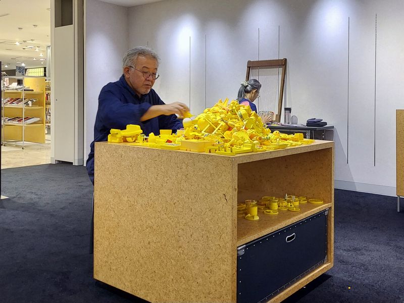 「まだどう展示するか決めてないんです」と、筆者と話しながら黄色のパーツを並べたり、崩したりすること十数分。「決めた!」とパーツを積み上げていく藤さん。壁側では、容子さんが、お菓子の袋を細く切って織りものを制作中。